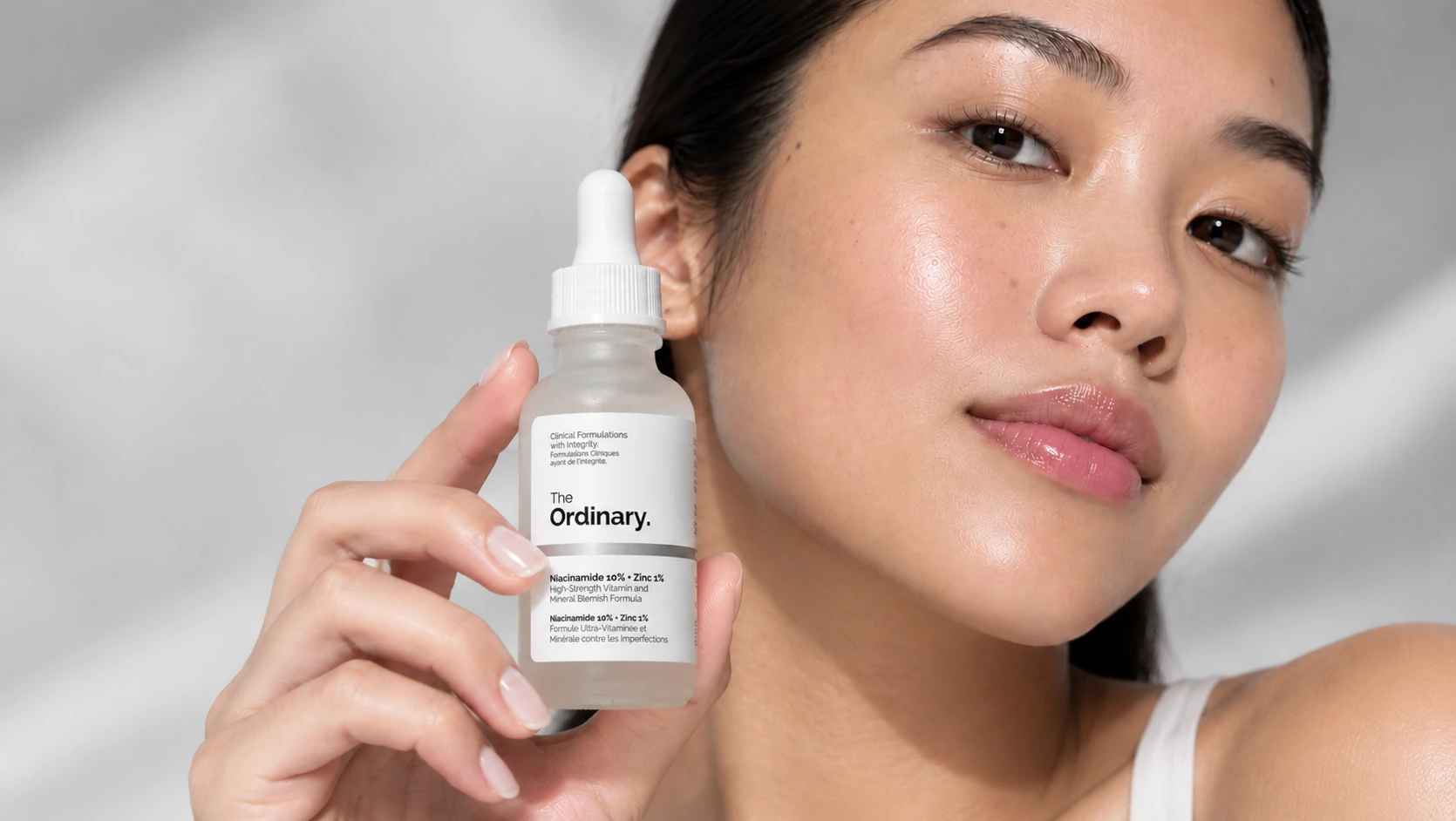Woman holding The Ordinary Niacinamide 10% + Zinc 1% serum beside her face